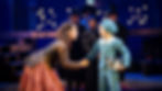 A woman leans toward a boy in a blue outfit holding books. Background shows people in dark clothes and top hats, set in a dimly lit stage.
