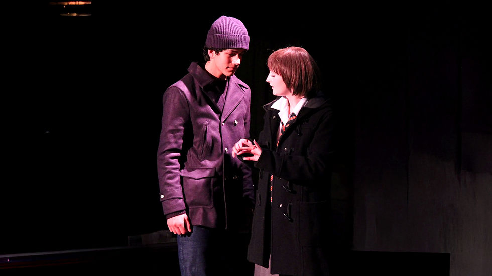 Two people in dark coats converse on stage in a dimly lit setting. One wears a beanie, the other has a bob haircut, conveying a serious mood.