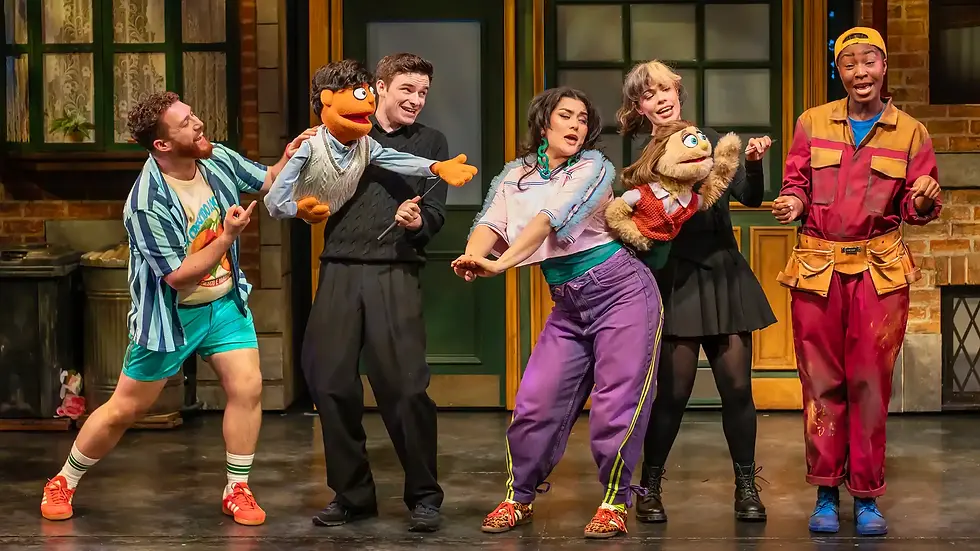 Performers on stage with two puppets, dancing energetically. Bright costumes and a colorful background evoke a lively, playful mood.