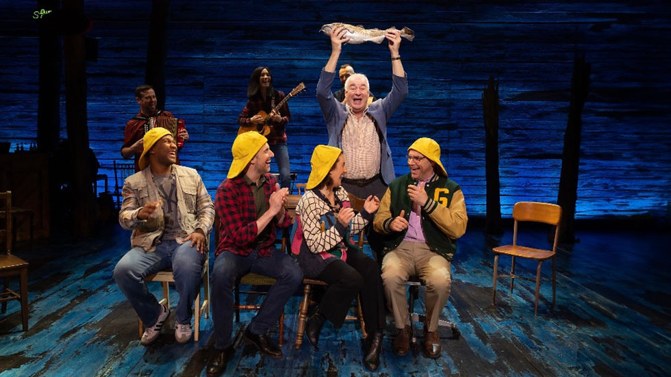 four people sat on wooden chairs on a stage with yellow hats on, whilst an old man with white hair holds a fish above his head with a happy expression. Taken from a bar scene in the musical Come From Away