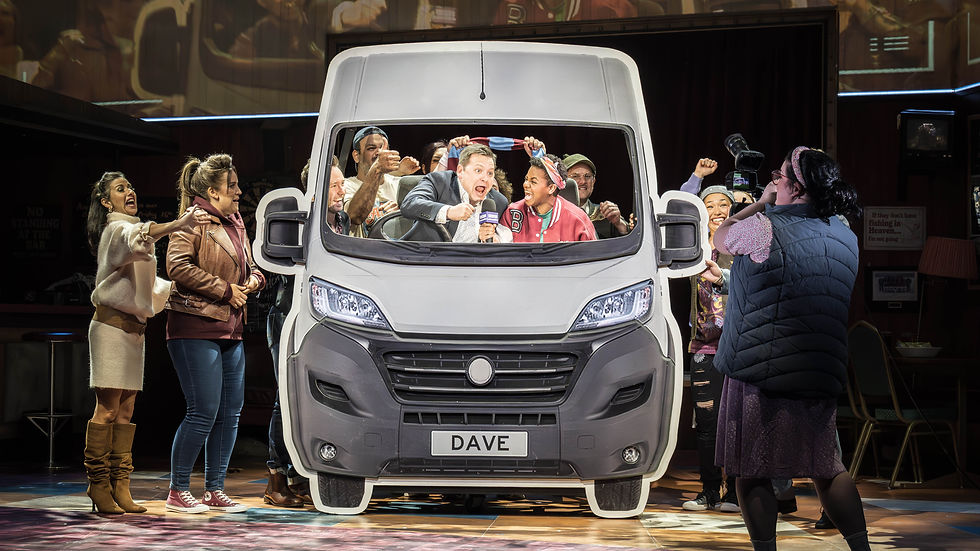 People cheer inside a van prop labeled "DAVE" on stage. A photographer captures the scene, evoking excitement and energetic mood.
