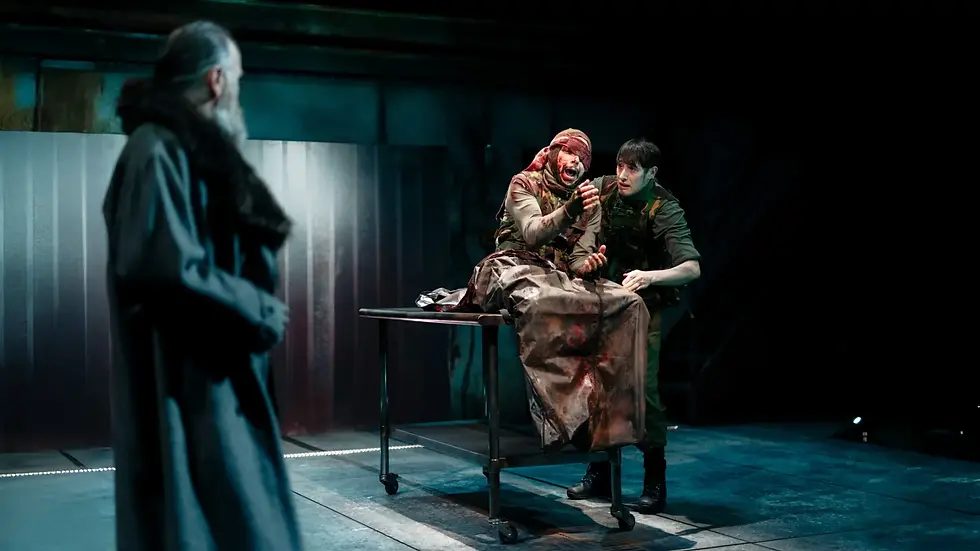 Two people in military gear sit on a metal table, one visibly distressed. A bearded figure in a coat looks on. Dim, industrial setting.