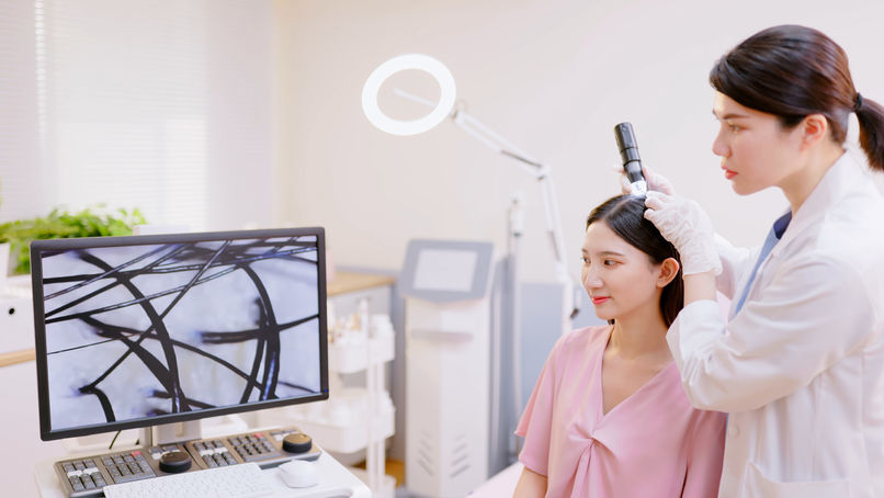A woman in a pink top receives a scalp examination from a person in a white coat using a device. Hair is shown magnified on a monitor.