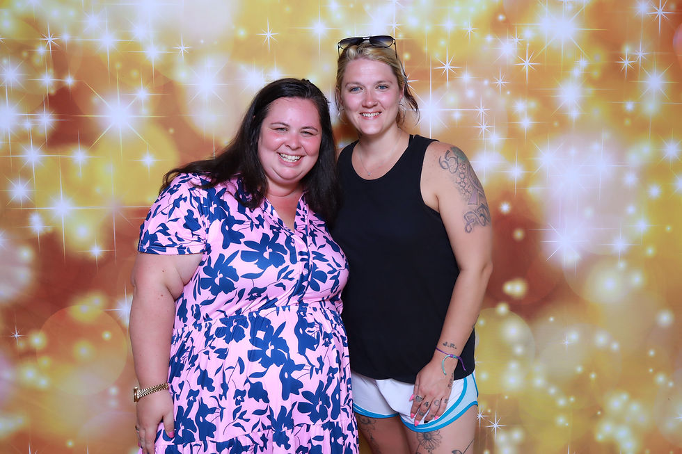 Two guests smiling in front of a sparkling gold photo booth backdrop during a group celebration.