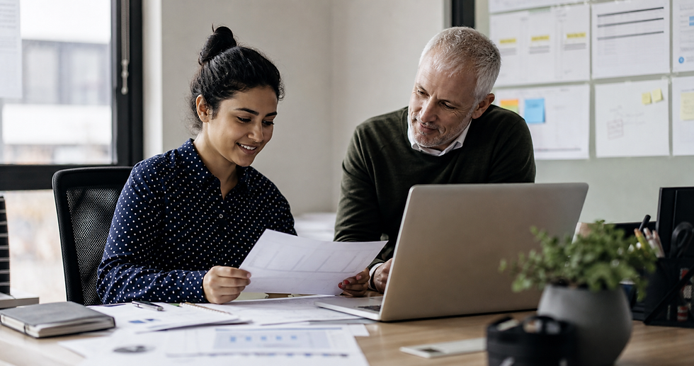 Two coworkers smile while reviewing a document at a laptop in a bright office with notes on the wall.