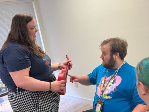 Music therapist holding a red ukulele while engaging a seated adult client during a session, with another observer nearby.