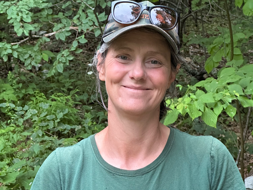 head shot of woman wearing a baseball cap, and a green tshirt in front of trees.
