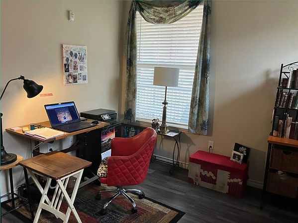 Part of a study with a wood floor, a large window, a writing desk, lamp and chair, along with a printer and bookshelves.