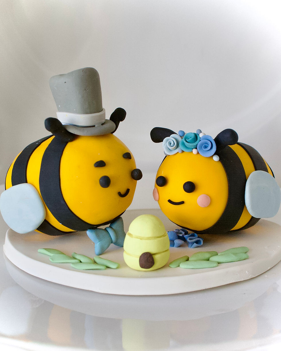 Two cute bee figures, one with a top hat and bow tie, the other with a floral crown. They're on a white base with green accents, smiling.