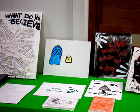 A collection of mental health awareness artwork displayed on a table with a vibrant green tablecloth. The display includes a canvas with collaged paper titled What Do You Believe, a simple illustration of blue and yellow figures titled Inner Child, a dark textured canvas with red lettering that reads Live with my brain for a day, and a small canvas depicting a lonely figure with text about trying like a lost child.