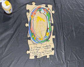 A top down view of a large wooden jigsaw puzzle piece resting on a black table cover. The center of the puzzle piece is painted with an expressive colorful oval ring of blue, green, and red strokes with a bright yellow explosion of color in the middle. Handwritten text at the bottom reads In all beings in all things and in all situations I will find the Light alongside the Latin phrase Credendo vides meaning By believing we see.