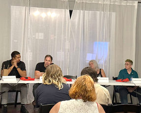 A closer view of the four person panel seated at the white folding table during the 2023 Fighting the Darkness event. The panelists are engaged, with one of the men speaking into a microphone. The backs of the audience members' heads are visible in the foreground.
