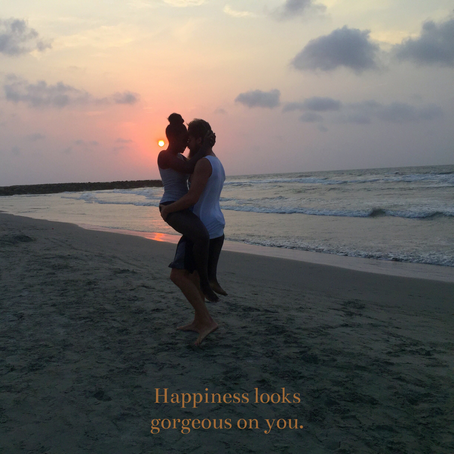 Couple embracing on a beach at sunset, warm sky hues, ocean waves. Text: "Happiness looks gorgeous on you." Romantic and serene mood.