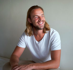 Dan The Happiness Coach smiling with long hair in a white shirt sits against a plain beige wall, exuding a cheerful mood.