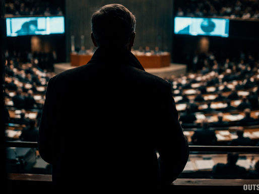 Homem solitário, visto de costas, observa um plenário político iluminado de cima, enquanto permanece nas sombras, destacando contraste entre lucidez individual e massa em movimento.