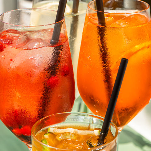 A close-up of long drinks in glasses with straws, showing rich orange and red tones, ice, and citrus garnish.