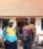 Two men, one in a racing shirt, stand before a tiled house
