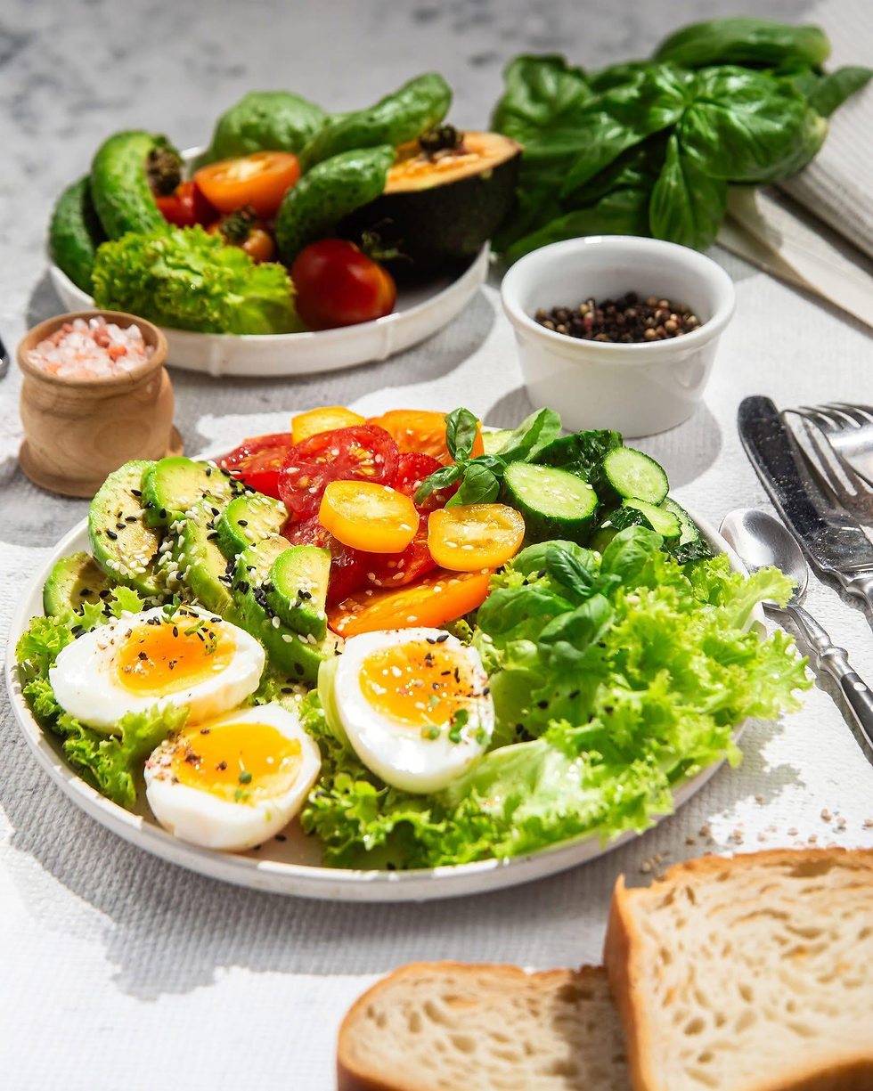 Đĩa salad rau xanh với bơ, trứng luộc, cà chua, dưa leo, bánh mì trên bàn sáng.