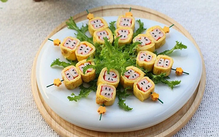 Món cuộn trứng kiểu sushi xếp thành bông hoa trên lá xanh, đặt trên đĩa trắng và khay gỗ, nền khăn sáng.