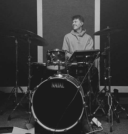 Jack Grange, a young man, smiling whilst playing an acoustic drum kit.
