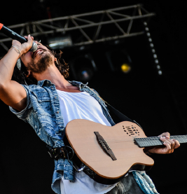 Bearded guitarist-singer belting into a mic onstage, wearing denim vest and white shirt, holding a tan acoustic guitar.