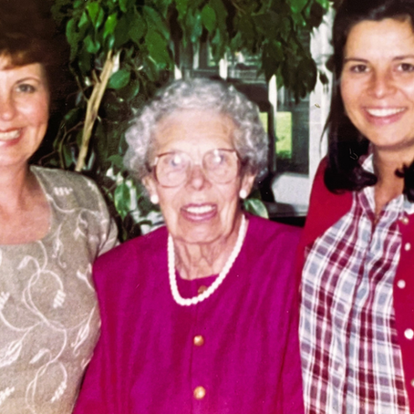 A family photo of three generations of women. Michele Barne's mother on the left in a patterned blouse, her grandmother in the center wearing a pink dress and pearls, and a younger Michele on the right in a plaid shirt and red cardigan.