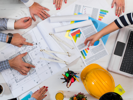 Overhead view of collaborative project planning meeting with multiple people's hands working on architectural blueprints, business charts, laptop, construction helmet, and measuring tools spread across a white table.