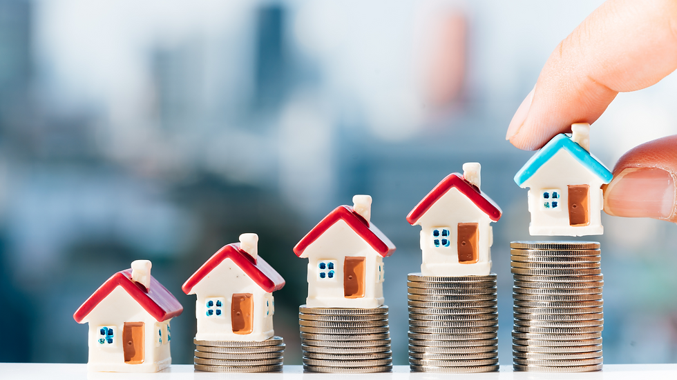 Toy houses on coin stacks show financial growth. A hand places a blue-roofed house on the tallest stack. Blurred cityscape backdrop.