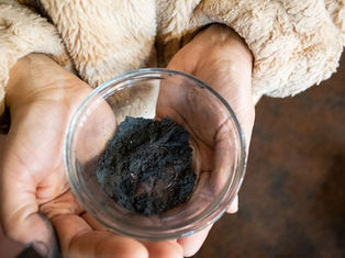 Manos sostienen un cuenco de vidrio con polvo negro. Fondo borroso. Abrigo de felpa beige. Sensación de curiosidad.