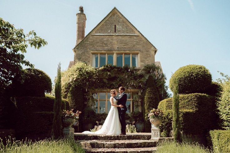 wide angle photo of bride and groom in a fancy garden with well-maintained hedges and a country house in the background.