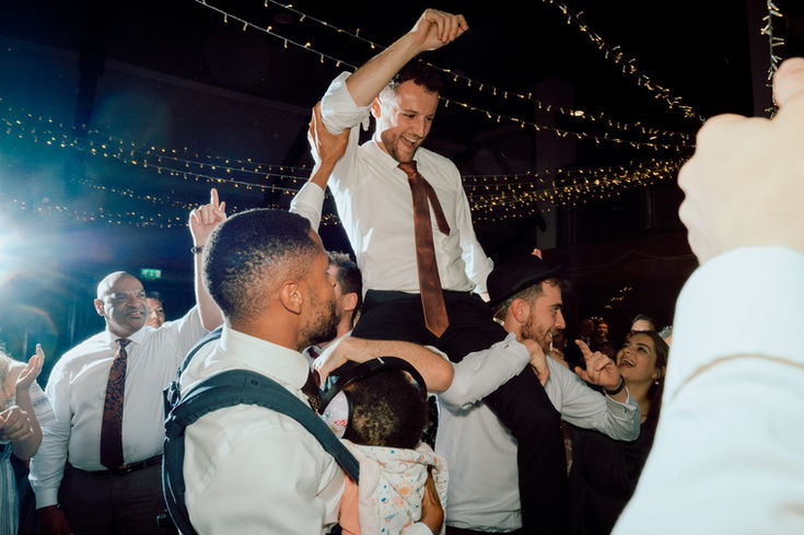 groom being lifted onto shoulders while on the dancefloor and surrounded by guests.