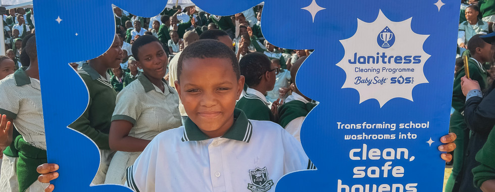 School children smile behind a blue splash photo frame during the Janitress Cleaning Programme event. They are celebrating with fellow learners gathered under an outdoor marquee.