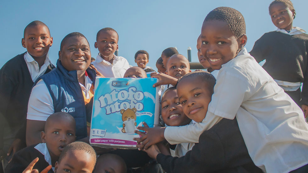 Smiling schoolchildren and WeSolve4X CEO, Tsietsi Ngobese, pose outdoors around a pack of Ntofo Ntofo toilet paper under a clear blue sky with new Amalooloo toilets in the background.