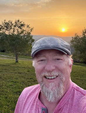 Ben Joslin smiling outdoors at sunset, wearing a flat cap and pink shirt, with trees and hills in the background.