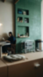 A person sits at a table using a smartphone in a cozy room with a green wall featuring shelves.