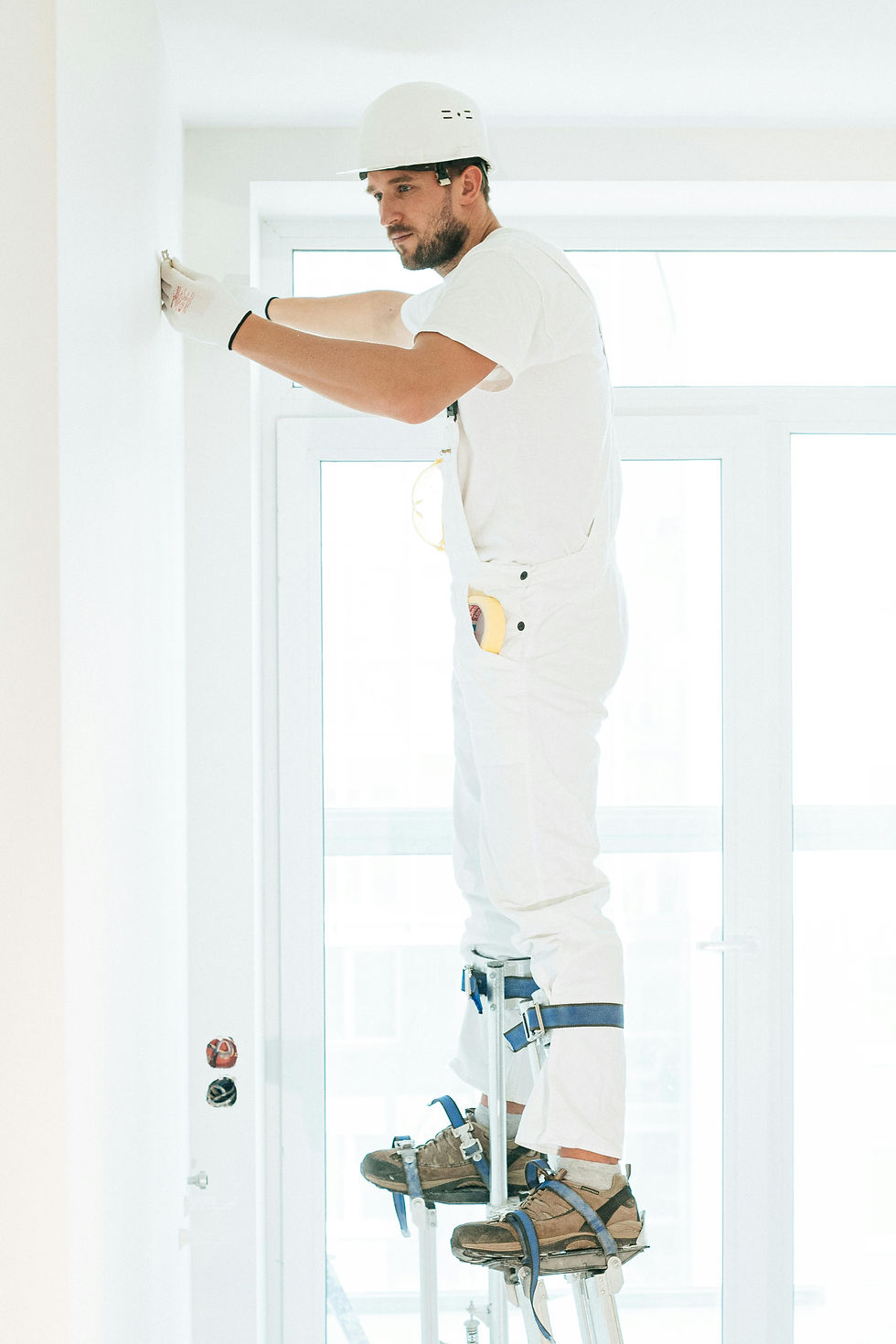 Worker in white outfit on stilts, adjusting wall fixtures. Bright room with large windows. Calm and focused expression.