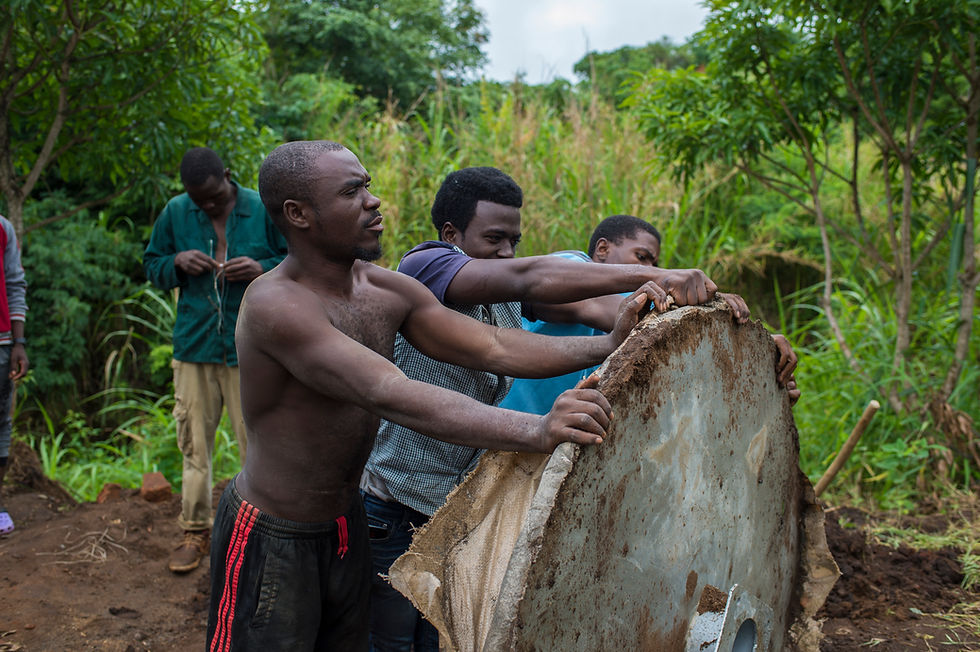 Three men push a large, round, water pump concrete base outdoors, surrounded by lush greenery. One man in the background works with wire.