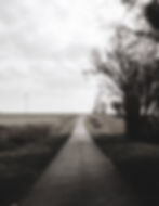 A one-lane country road with flat fields on either side in black and white