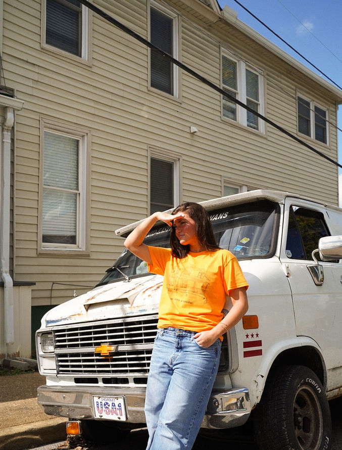 Woman wearing The Fort Volicious T-shirt standing beside a white Chevrolet van in sunlight