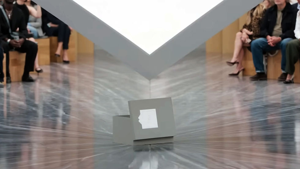 A gray box with a cut-out face pattern on the floor surrounded by seated audience in a reflective, modern space. Bright lighting overhead.