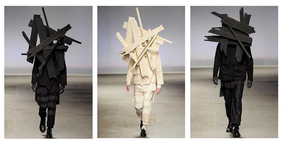 Models on a runway wear black and white outfits with oversized wooden planks. The setting is minimal, with a neutral background.