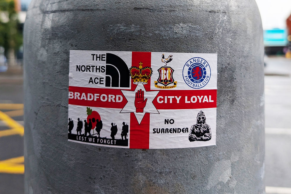 Sticker on a pole with "Bradford City Loyal" text, featuring logos, a red hand, crown, and silhouettes. Background shows a street scene.