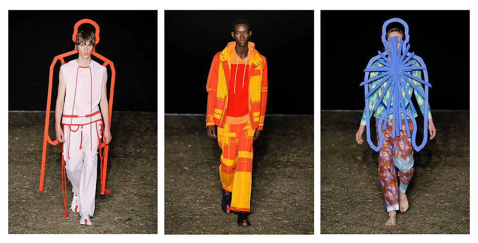 Three models in colorful, avant-garde outfits walk a runway. Background is dark; outfits include vibrant orange, red, blue, and abstract patterns.