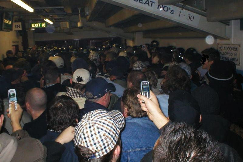Crowd in a stadium corridor faces police. People hold up phones. Signs read "EXIT B" and "NO ALCOHOL ALLOWED." Tense atmosphere.