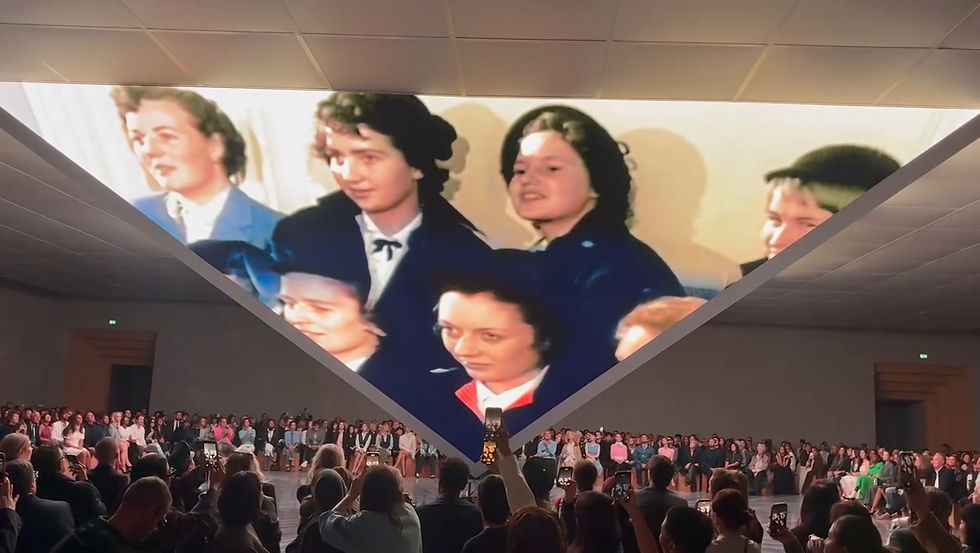 Large screen with vintage video of women in blue coats, surrounded by crowd in a modern venue. Mood is nostalgic and engaging.