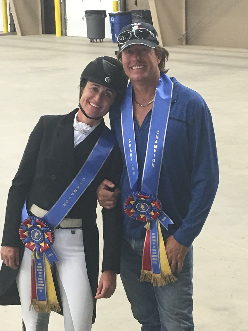 Lauren Ogden and Tim Forster smiling, wearing blue "Champion" sashes with rosettes. Indoor setting with concrete floor and trash bins in the background.