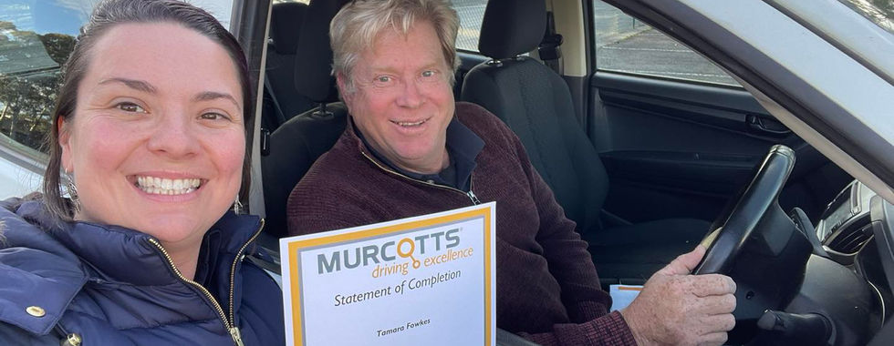 Rotarian Tamara Fowkes standing outside a silver car holding up a murcotts certificate. Fellow rotarian Chris is sitting in the drivers seat of the car, smiling out the window.
