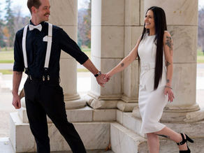 Smiling couple holding hands between marble columns, he in black with suspenders, she in white dress and heels.
