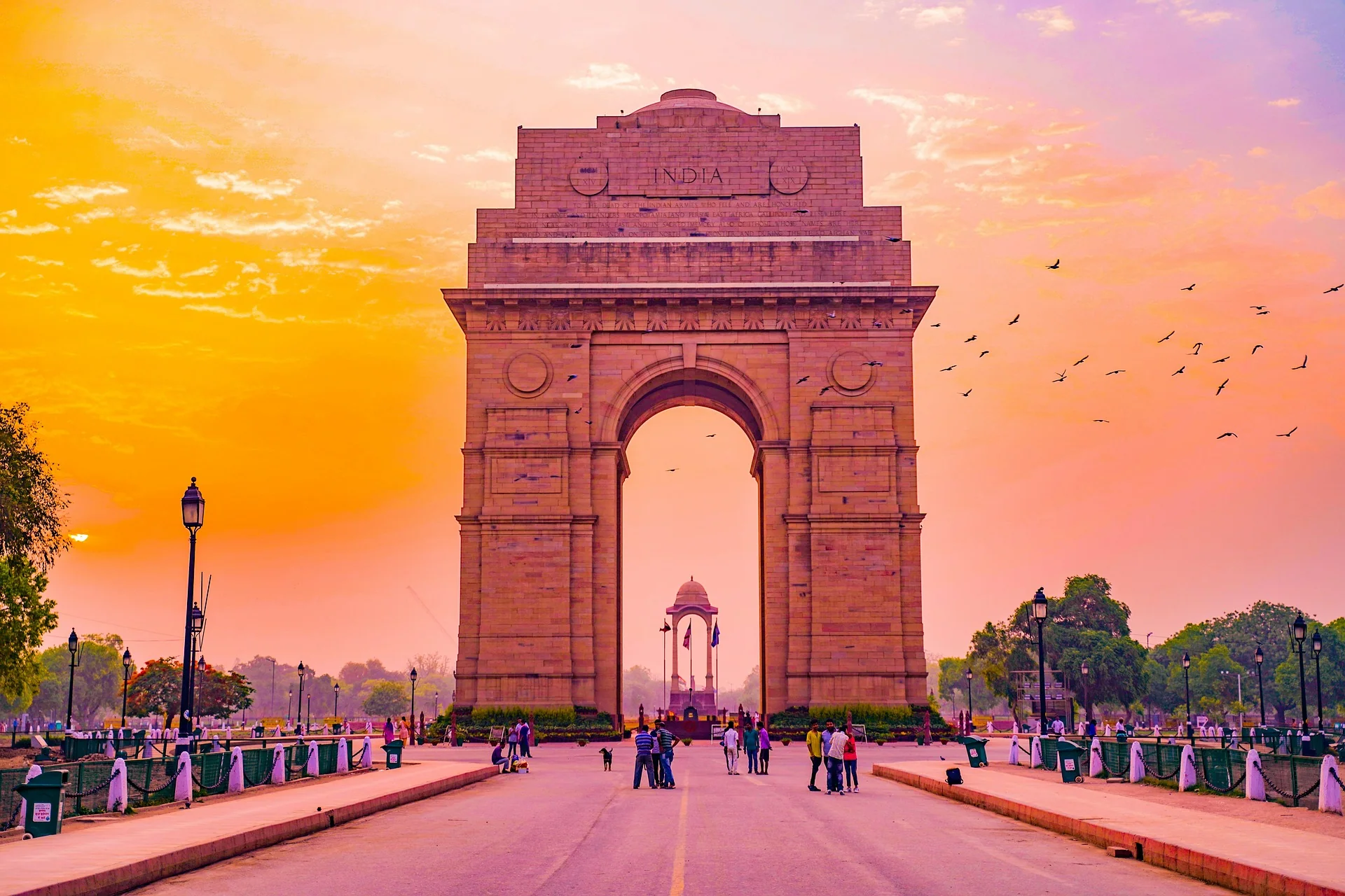 A large sandstone arch monument with the words "INDIA GATE" engraved on the top, surrounded by a circular lawn and a fountain. The monument is lit up by spotlights and has the Indian flag flying on both sides.
