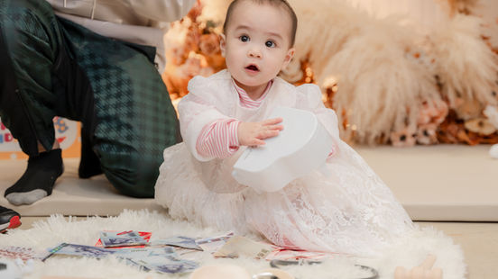 寶寶身穿白紗裙坐於地毯上,手握抓周道具專注凝視,背景奶茶色乾燥花藝柔美綻放,氛圍細膩溫馨